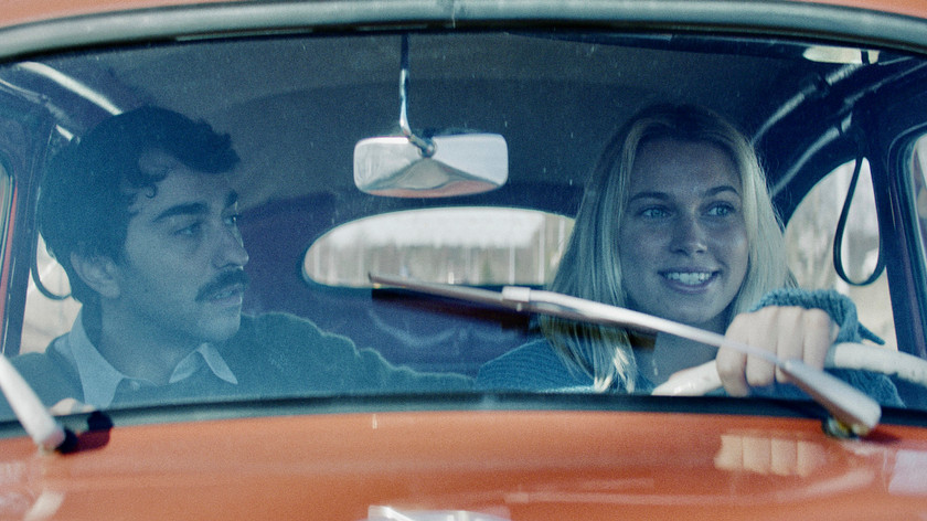 Gjennom frontruten på en oransje bil ses en mann med mørkt hår og bart, og en dame med lyst langt hår. Damen sitter bak rattet. Foto.