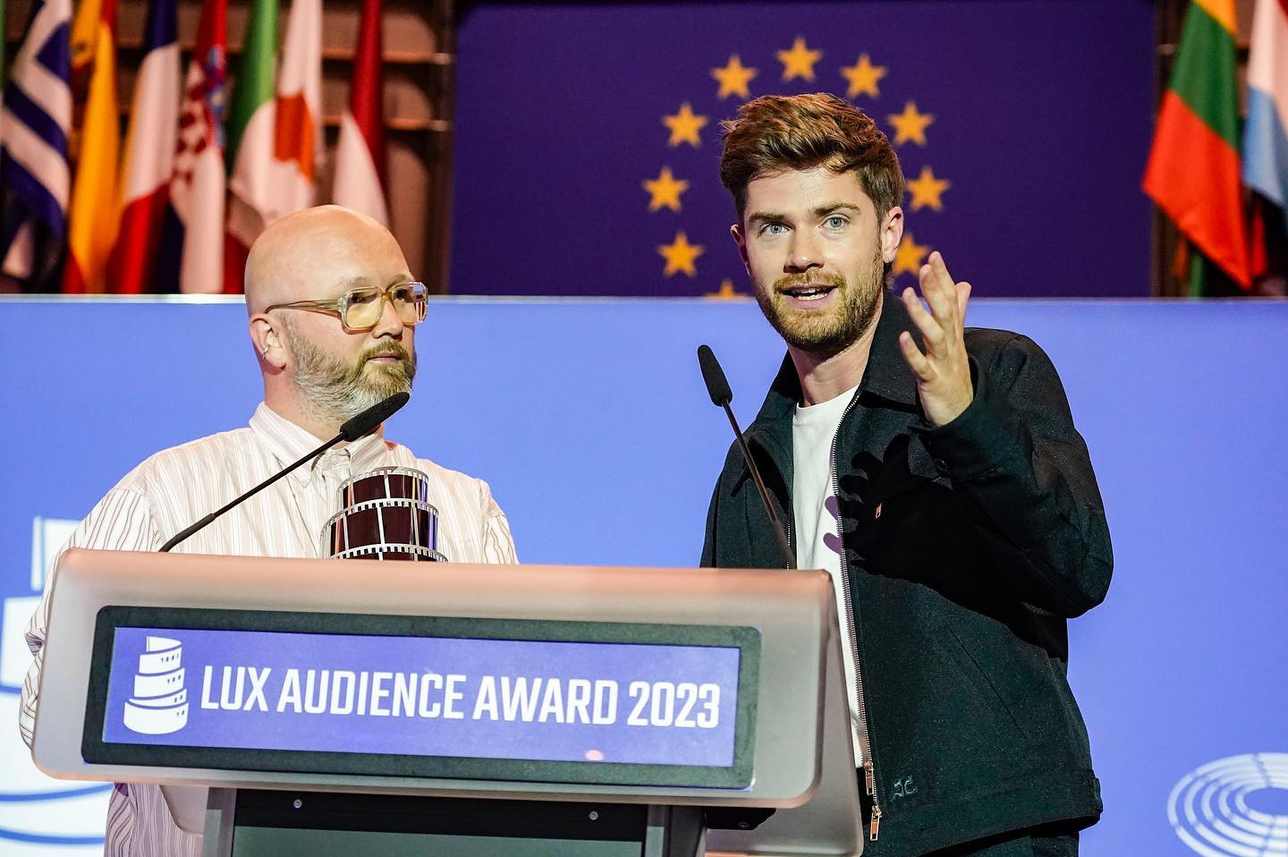 Lux Audience Awards 2023. To menn står bak en talerstol. Den ene holder en statuett i sølv formet som en filmrull, den andre snakker i mikrofonen. I bakgrunnen skimtes EU-flagget og en rekke andre flagg. Foto.