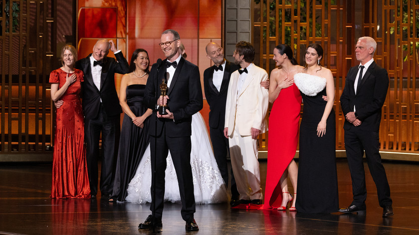 Foto. En gruppe festkledde og glade personer på en scene. En mann i smoking holder en statuett.