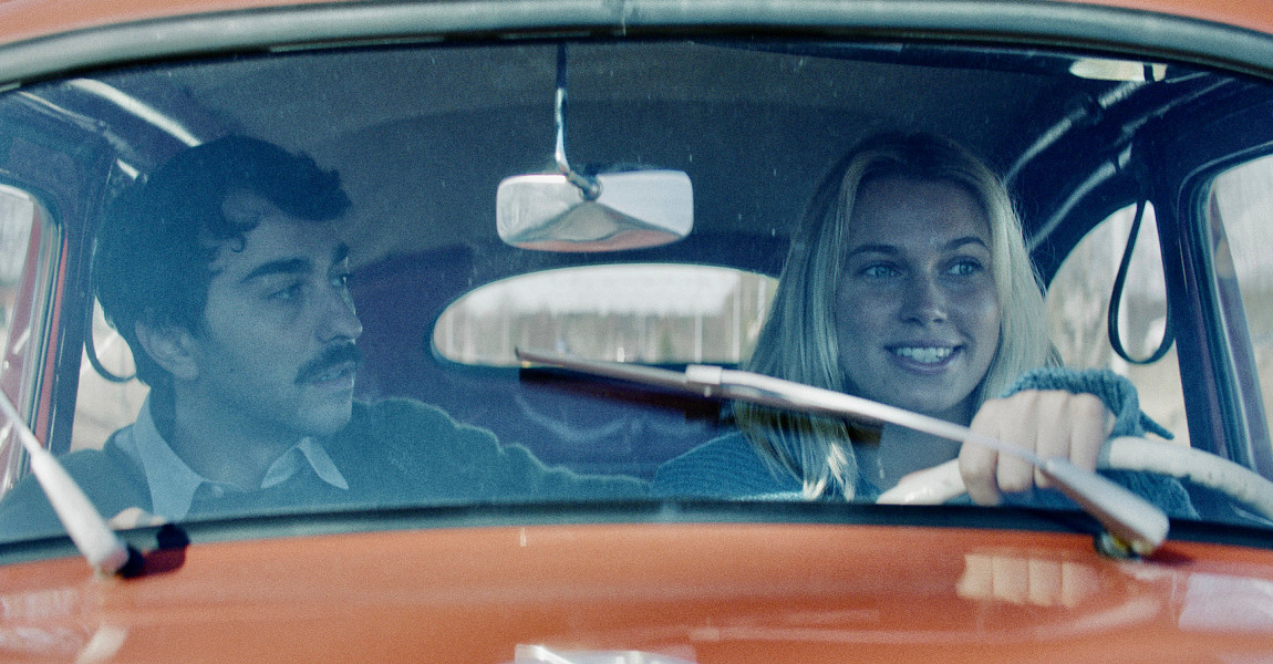 Gjennom frontruten på en oransje bil ses en mann med mørkt hår og bart, og en dame med lyst langt hår. Damen sitter bak rattet. Foto.