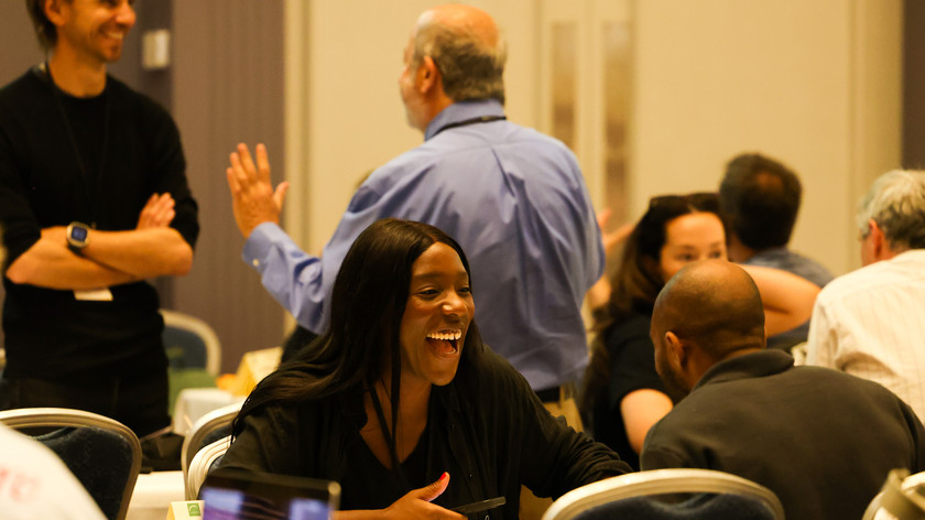 Foto. Flere personer i et rom. De sitter to og to og prater sammen ved bord.