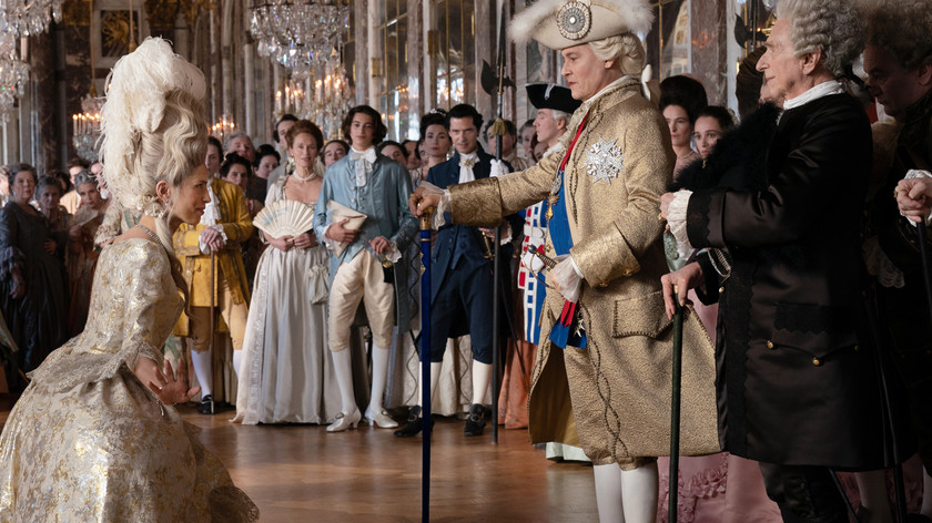 En gruppe mennesker i 1700-talls festklær er samlet i en staselig ballsal. I forgrunnen neier en dame foran en mann. Foto.