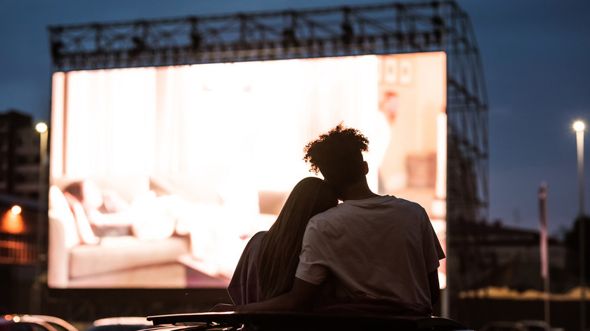 Et ungt par, ei jente og en gutt, er på drive-in kino utendørs. De holder rundt hverandre. Det er kveld, de sitter med ryggen til oppå en bil. Foran dem vises en film på stort lerret og rundt står flere andre biler parkert. Foto.