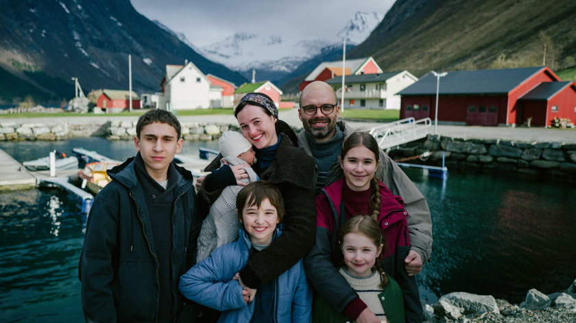 Foto. En familie med to voksne og fem barn står ved et vann med fjell i bakgrunnen. 