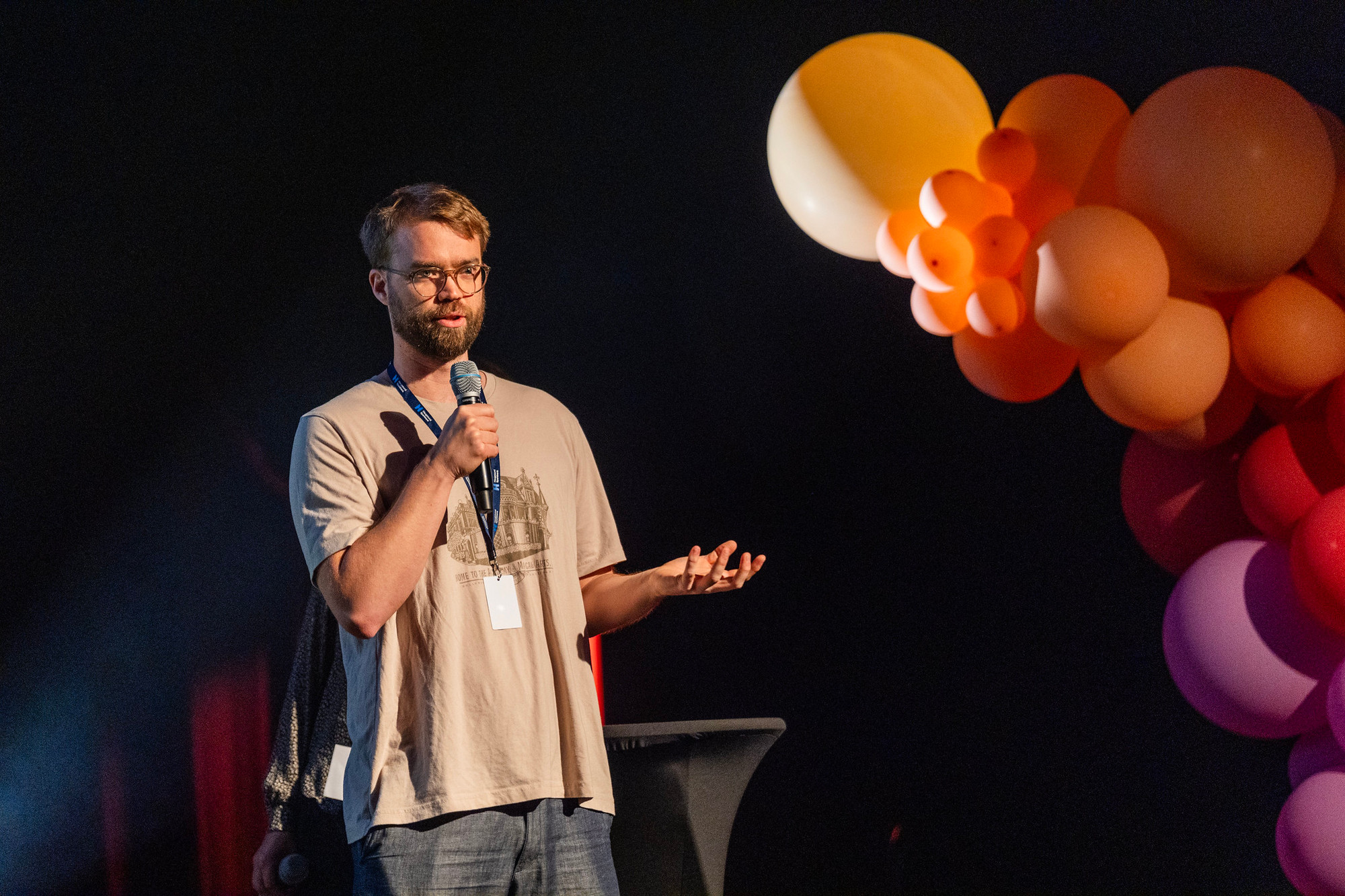 Foto. En mann står på en scene og snakker i en mikrofon. Ballonger i bakgrunnen.