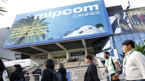 Flere store plakater henger over et inngangsparti. Den ene er en reklame for mipcom Cannes. Flere personer går forbi nedenfor inngangen. Foto.