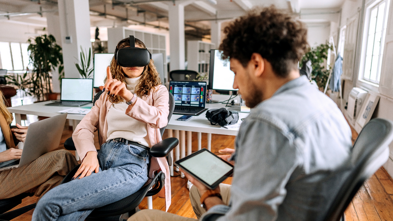 En dame og en mann sitter i et åpent kontorlandskap. Hun har på seg VR-briller og peker opp i luften, han sitter med et skjermbrett foran seg. Foto.
