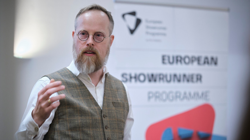 En mann med briller og skjegg, ikledd skjorte og rutete vest foreleser foran en plakat med teksten European Showrunner Programme. Foto.