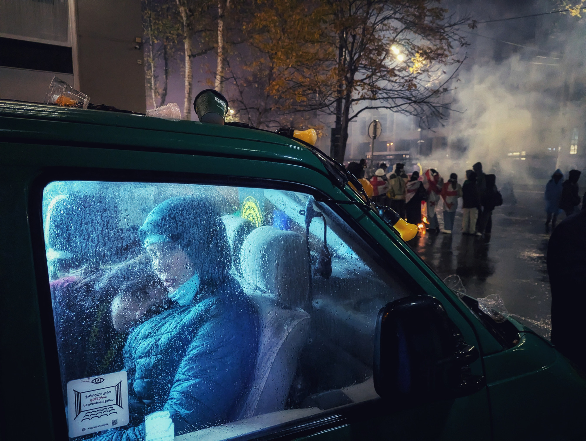 Foto. Et par kvinner skimtes gjennom et bilvindu. I bakgrunnen ses røyk og demonstranter i en mørk gate.