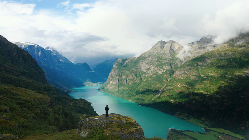 En mann står og ser ut over et storslått landskap med fjell og fjor. Foto.