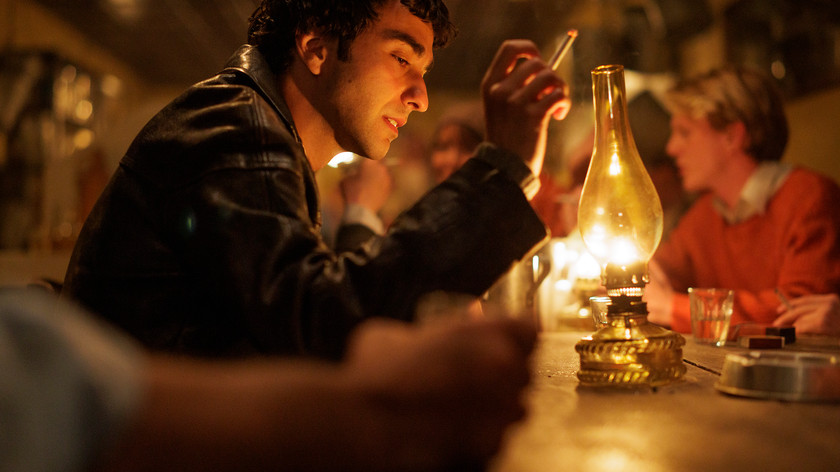 En mann sitter ved en bardisk i lyset fra en oljelampe. Han holder en sigarett i hånden. Flere andre skimtes i bakgrunnen. Foto.