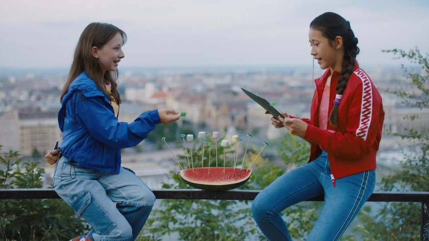 Foto. To jenter sitter på et gjerde med utsikt over byen. Den har en kvart vannmelon mellom seg. I den er det stukket pinner med smågodt på enden. Den ene jenta ser på et ark. Den andre smiler og ser på henne.