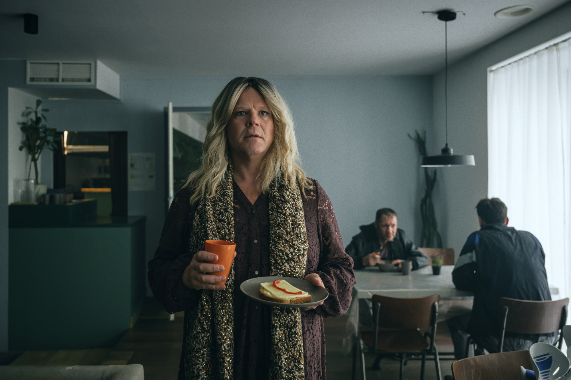 Foto. En mann i kvinneklær holder en asjett med ei brødskive og en kopp. I bakgrunnen sitter to menn ved et bord.