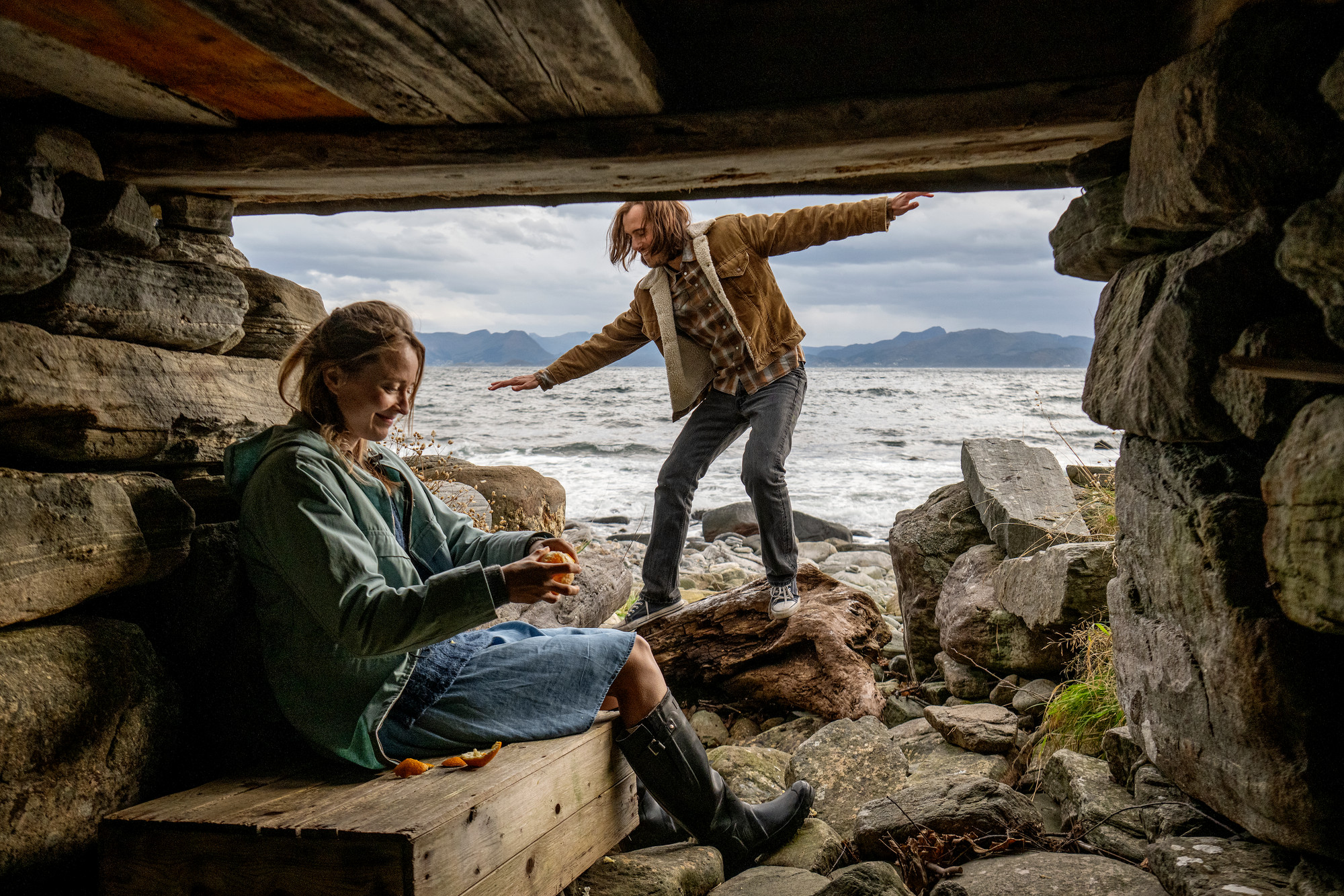 Foto. Ei dame sitter under et overbygg i fjæra og skreller en appelsin. En mann balanserer på en stein utenfor.