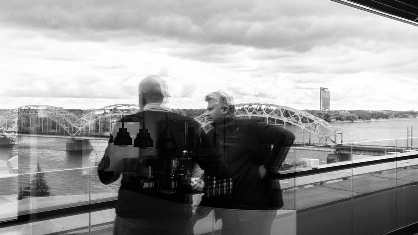 To menn står ute på en balkong med utsikt over en elv. Over elven går en bro. Bebyggelse skimtes på den andre siden. Foto.