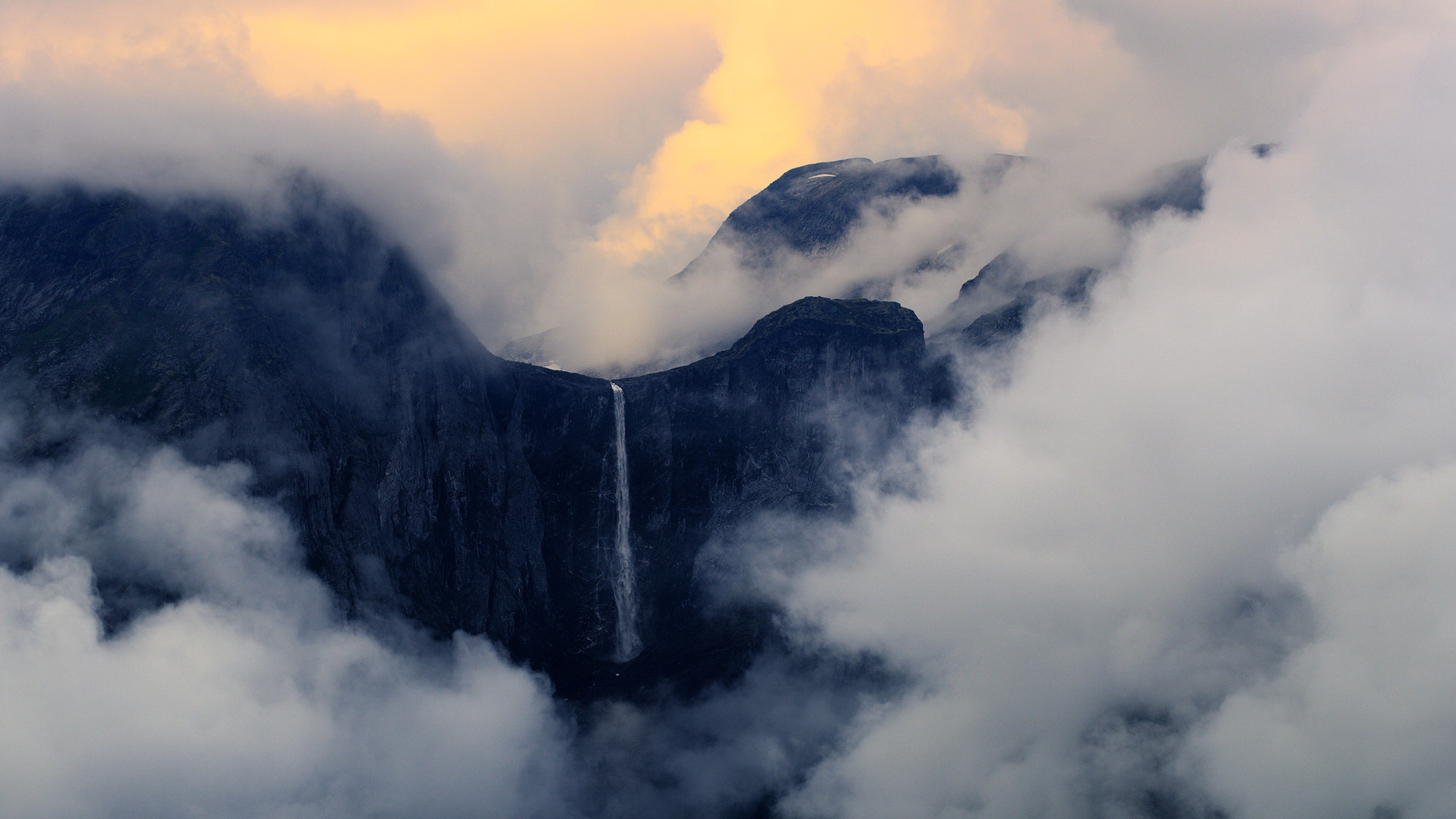 Fjellandskap med fossefall. Skyer omkranser fjellene. Foto.