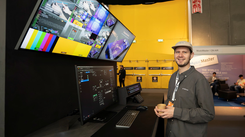 Foto. En mann med caps smiler mot kamera. Han holder en pappkopp og står ved siden av flere skjermer.