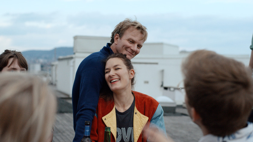 En mann holder rundt en kvinne, begge smiler. De sitter ute på et tak sammen med en gruppe mennesker. Foto.