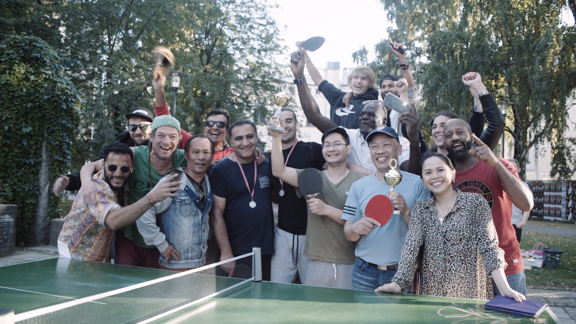 En gjeng med jublende personer står bak et bordtennis-bord. De holder rekkerter og en pokal. Foto.