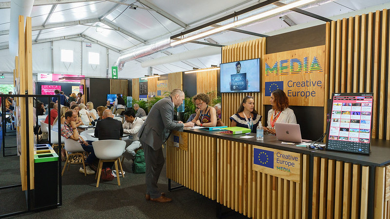 Foto. En stand inne i et telt. Flere personer står ved en disk og sitter ved runde bord på området.