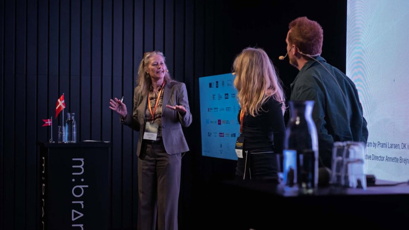 Foto. Tre personer på en scene. To har ryggen vendt til. En dame står bak en talerstol og prater og ser mot de andre. Flere skjermer skimtes i bakgrunnen.