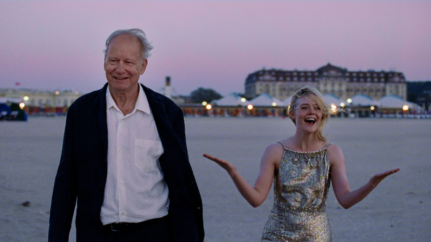 Foto. En eldre mann og en ung dame i pentøy på en strand med et flott hotell og partytelt i bakgrunnen. Begge smiler.