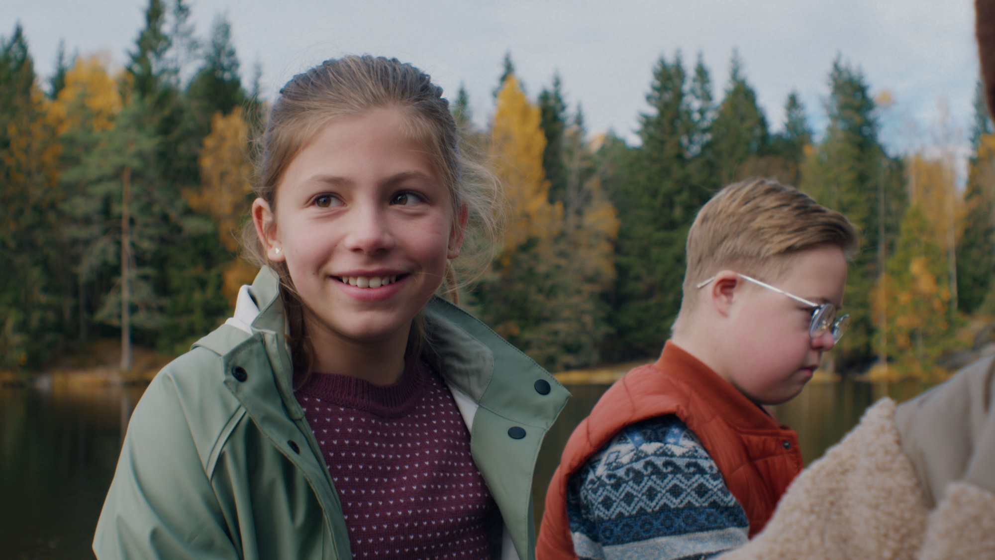 Foto. Ei jente og en gutt står ved et vann med skog i bakgrunnen. 