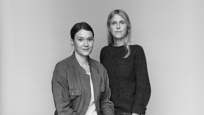Foto. Portrettfoto i sort hvitt av to kvinner. Den ene står og den andre sitter.