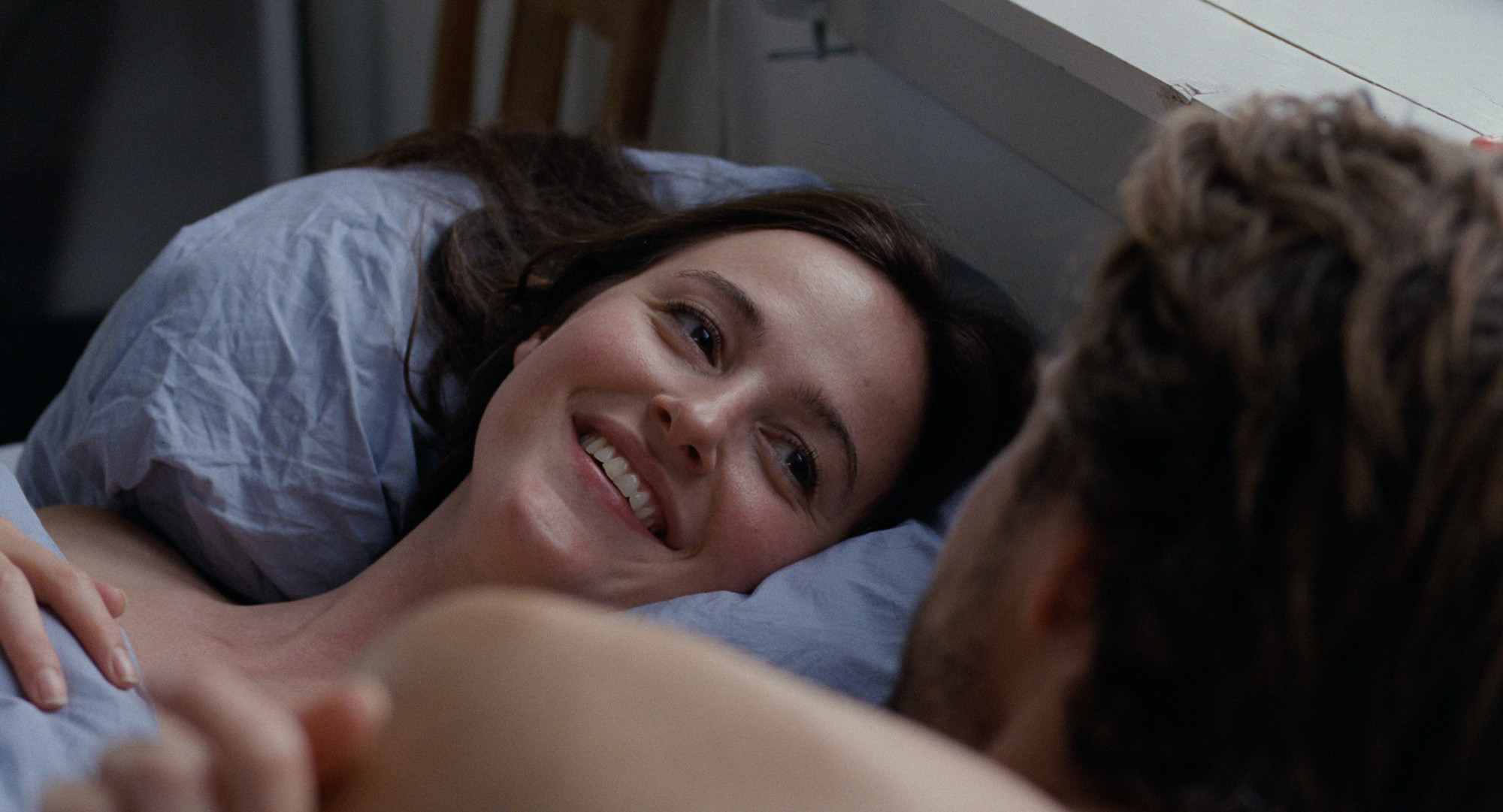 Foto. En kvinne ligger i sengen og smiler mot en mann.