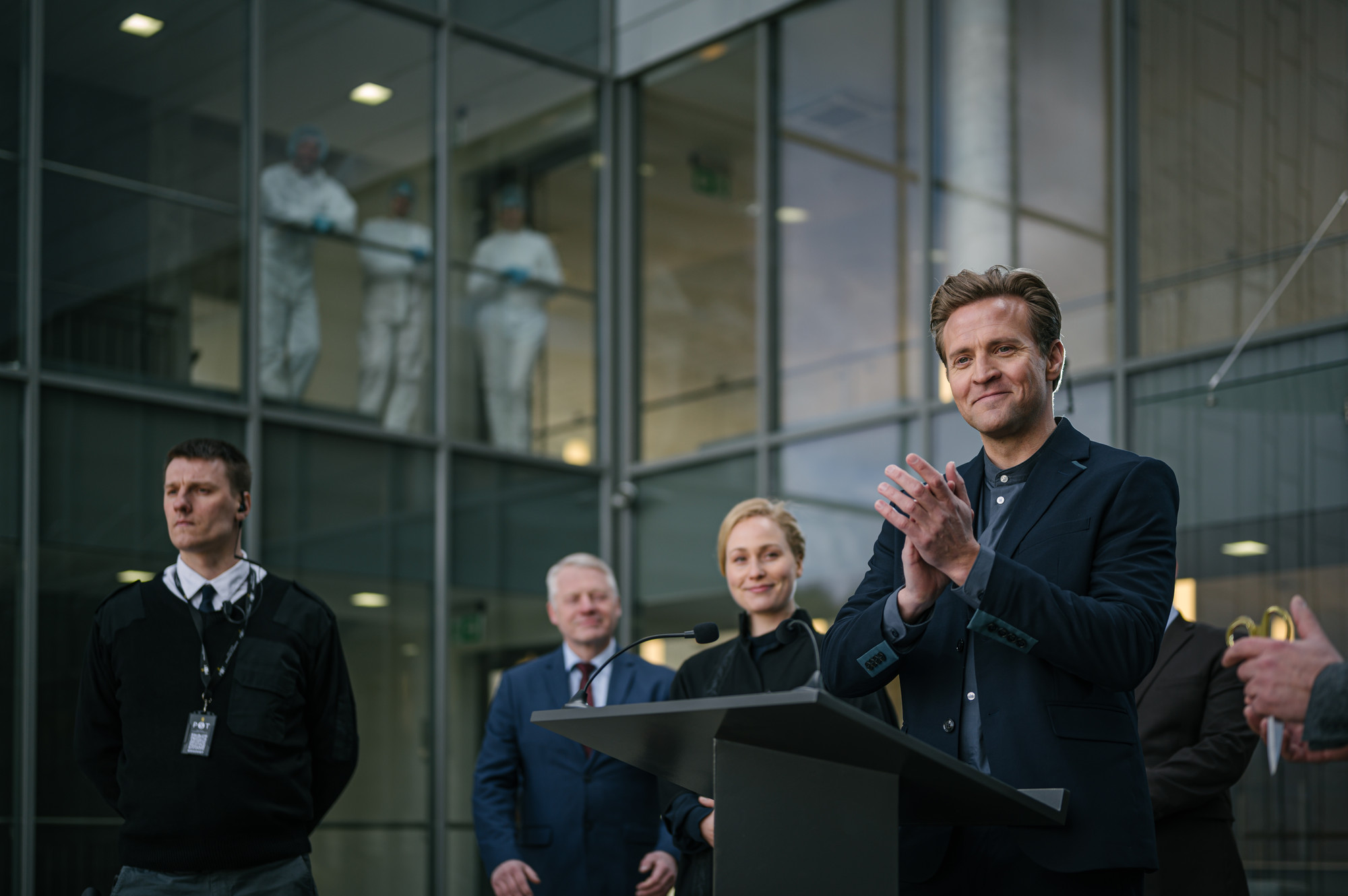 En mann med blondt kort hår og dress står bak en talerstol. Han smiler og klapper. Rundt han i bakgrunnen står flere personer, bl.a. en sikkerhetsvakt. I glassbygget i bakgrunnen skimtes tre personer med hvite drakter og blå nett på hodet og hansker på hendene i et vindu i 2. etg..