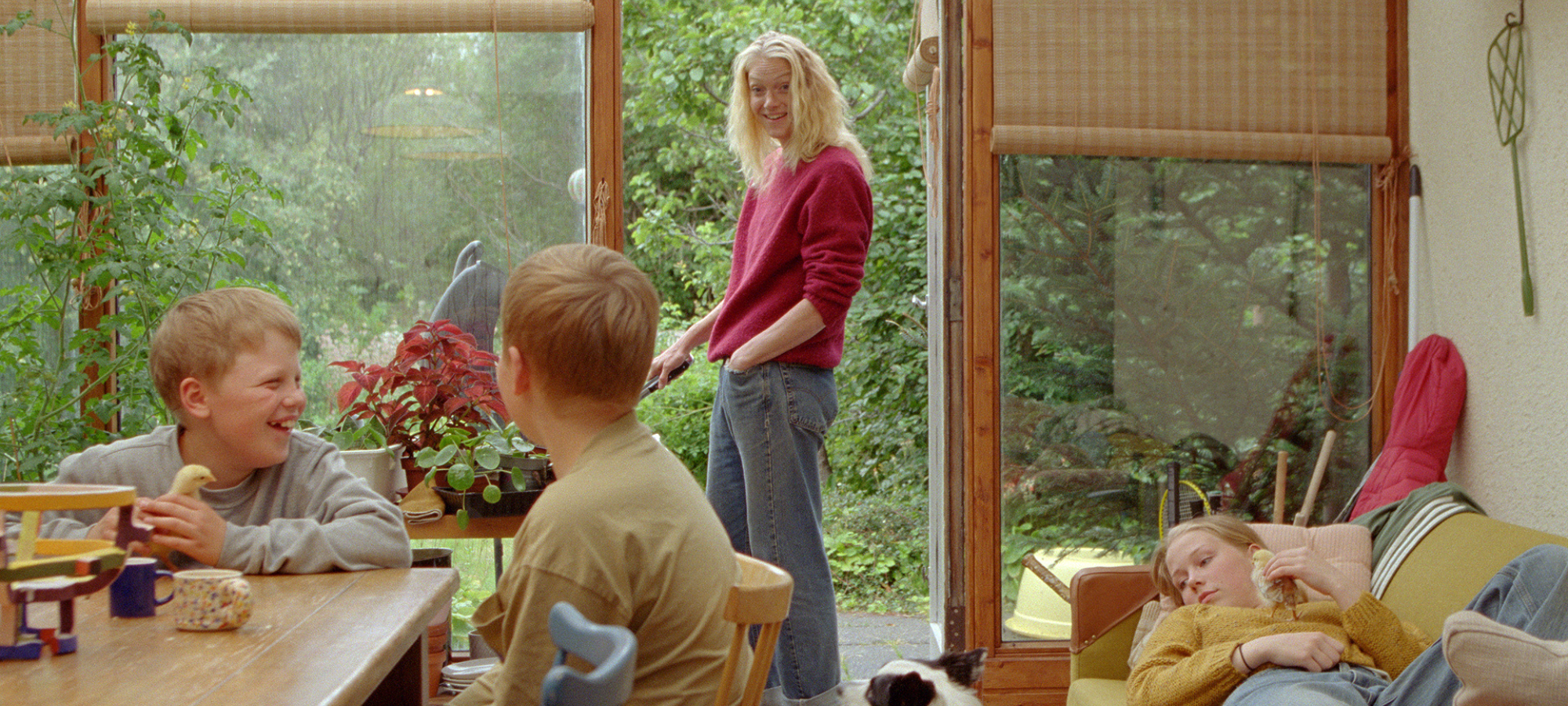 Foto. En dame står i en døråpning ut til en hage. Innenfor døra sitter to gutter ved et bord og ei jente ligger på en sofa. Den ene gutten og jenta holder hver sin gule kylling.